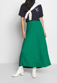 Groene maxi-rok gemaakt van lichtgewicht stof, gecombineerd met een marineblauwe t-shirt en witte hoge laarzen. Opvallende textuur en snitdetails zichtbaar.