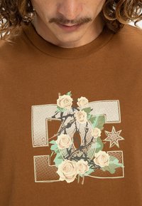 Braunes Baumwoll-T-Shirt mit einem großen Grafikdesign, das Rosen, Äste und einen Stern zeigt, umrissen in einer hellen Farbe mit gepunkteten Akzenten.