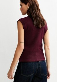Vrouw met bruin haar in een mouwloos kastanjebruin gebreid topje en donkerblauwe jeans, staande zijwaarts tegen een witte muur.