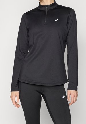 Kobieta ubrana w czarną, długorękawową sportową bluzę na zamek oraz dopasowane legginsy, oba z białym logo ASICS po lewej stronie.