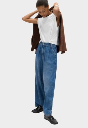 Kvinde iført løstsiddende blå jeans, hvid ærmeløs top, sorte sko, iført en brun jakke mod en ensfarvet lys baggrund.