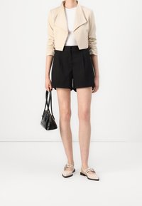Beige cropped jakke med opsmøgede ærmer, hvid tætsiddende top, sorte skræddersyede shorts og beige slip-on sko; bærer en lille sort håndtaske.