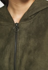 Olivgrön, zip-up jacka med huva, tillverkad av mjukt, strukturerat material. Har en synlig dragkedja och ringdragning.