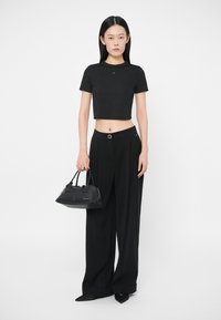 Γυναίκα με μαύρο κοντό μπλουζάκι, φαρδιά μαύρα παντελόνια και μαύρες μυτερές γόβες, κρατώντας μια μαύρη τσάντα μπροστά σε λευκό φόντο.