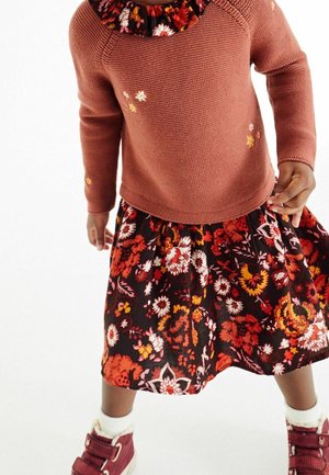 Enfant portant un pull en tricot couleur rouille avec broderie florale, une jupe noire à motifs floraux, des chaussettes blanches et des chaussures bordeaux.