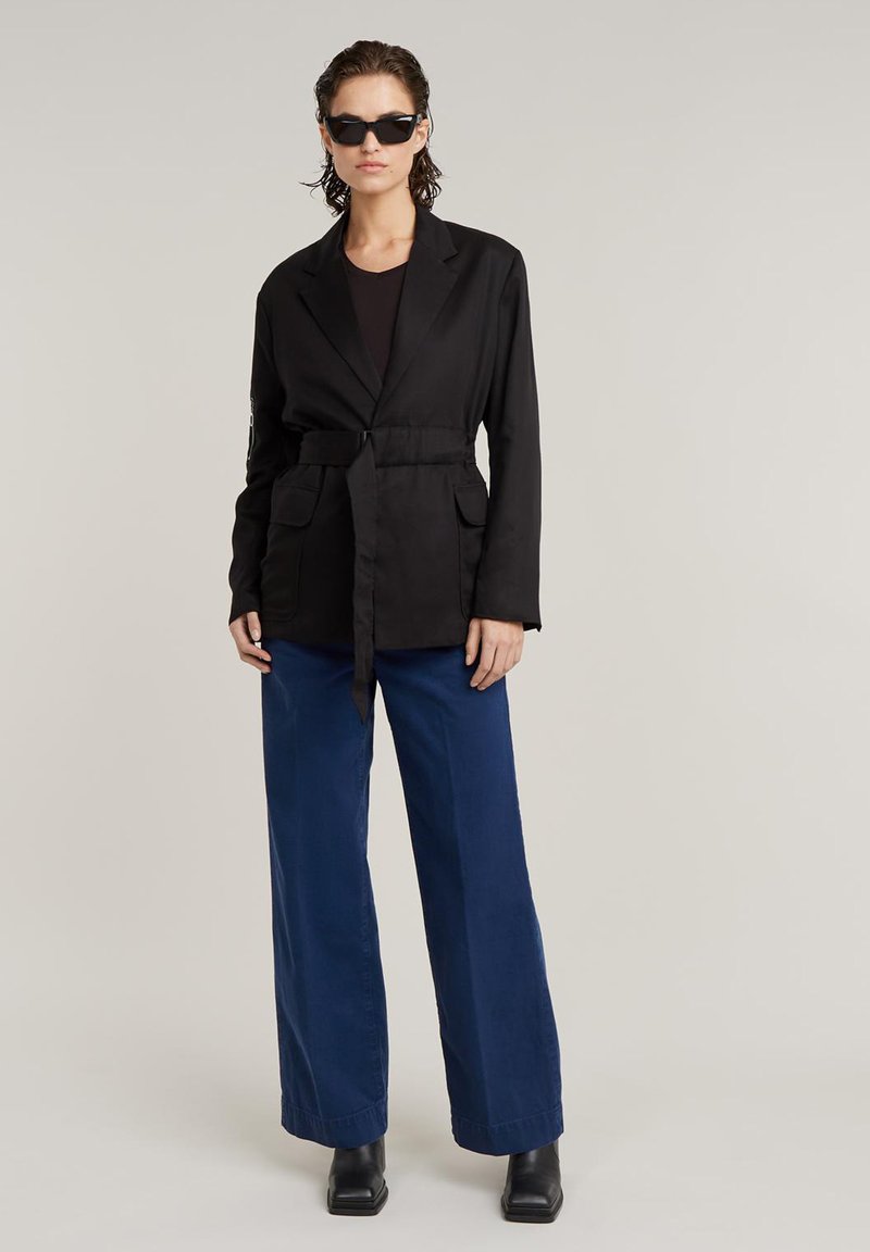 Kobieta ubrana w czarny żakiet z paskiem, czarną bluzkę, niebieskie szerokie spodnie, czarne botki na kwadratowym obcasie oraz czarne okulary przeciwsłoneczne, stojąca na jasnym tle.