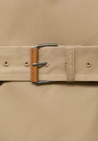 Beige stof met een brede riem met een zilveren metalen gesp en een bruine leren accent. Stiknaden zichtbaar langs de randen van de riem.