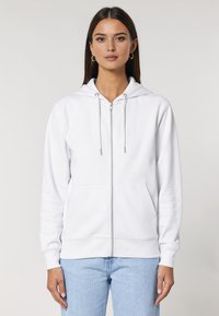 Felpa bianca con zip realizzata in tessuto morbido, dotata di cappuccio, lacci e tasche anteriori. Manicotti e orlo a costine, abbinata a jeans azzurri chiari.