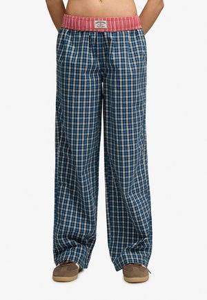 Personne portant un pantalon large à carreaux bleu et blanc avec une ceinture rouge et des chaussures décontractées beiges, debout devant un fond uni.