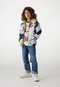 Giacca imbottita con una stampa grafica multicolore, accenti verdi e cappuccio, abbinata a una maglietta bianca e jeans in denim blu, sneakers bianche.