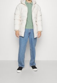 Hvid pufferjakke med hætte, der har lynlås og knaplukning, båret over en grøn polo skjorte og blå jeans, afsluttet med sorte sneakers.