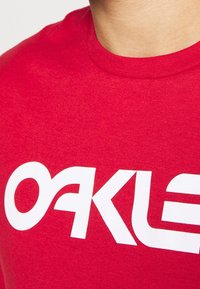 Czerwona bawełniana koszulka z dużym białym logo "OAKLEY". Materiał ma gładką fakturę, a koszulka posiada okrągły dekolt.
