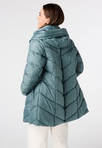 Vrouw die glimlacht, gekleed in een teal kleurige gewatteerde pufferjas met capuchon, staande met handen in de zakken, met de zijkant naar een witte achtergrond gericht.