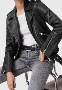 Persoon in een zwarte leren jas, wit T-shirt, grijze high-waisted spijkerbroek en zwarte riem die op een chroomstoel zit.