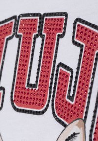 Weißes T-Shirt mit großen, rot glitzernden Buchstaben, umrandet in Schwarz, und einem Grafik-Design eines Cartoon-Gesichts in der unteren Ecke.