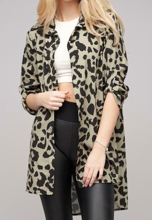 Femme portant une longue chemise boutonnée verte à imprimé léopard sur un crop top blanc et un legging noir.