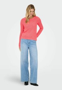 Femme portant un pull en maille corail, un jean bleu clair à jambes larges et des bottines noires à talons, debout avec une main derrière le dos.