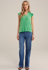 Top verde a maniche corte con scollo a V e dettagli arricciati, abbinato a jeans blu a campana. Il tessuto sembra liscio e leggero.