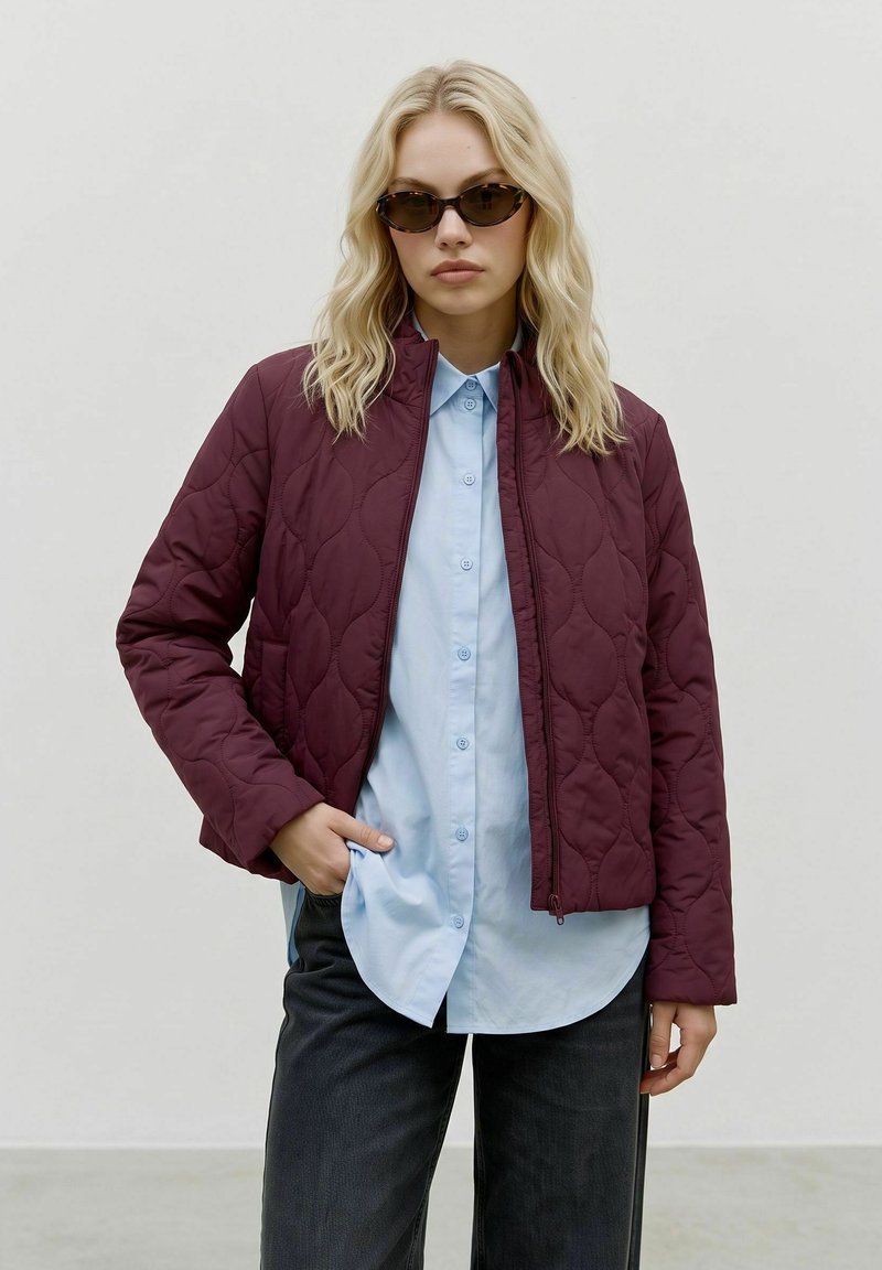 Femme blonde portant des lunettes de soleil foncées, une veste matelassée bordeaux, une chemise bleu clair boutonnée et un pantalon noir, debout avec une main dans la poche.