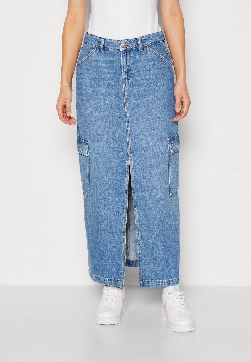 Femme portant une jupe droite en jean bleu avec une fente frontale et des poches latérales, associée à des baskets blanches et un top blanc.