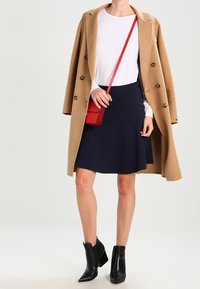 Cappotto cammello, camicia bianca a maniche lunghe, gonna svasata blu navy, stivaletti neri alla caviglia, borsa rossa a tracolla. Texture lisce e colori a contrasto.