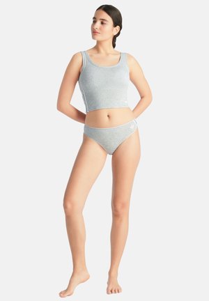 Frau mit dunklen Haaren im Pferdeschwanz, die ein graues ärmelloses Kurztop und passende niedrig geschnittene Unterwäsche trägt, barfuß auf einem schlichten Hintergrund stehend.