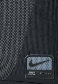 Le produit Nike Utility 2.0 présente un maillage noir texturé et un tissu durable, avec une étiquette de logo ovale proéminente en gris clair et des accents noirs.