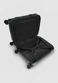 Valigia rigida nera con quattro ruote; l'apertura con cerniera mostra un interno vuoto con cinturini incrociati e una tasca in rete.