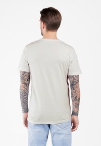 Hombre con tatuajes en ambos brazos, vistiendo una camiseta beige clara y jeans azules claros, de espaldas sobre un fondo blanco.