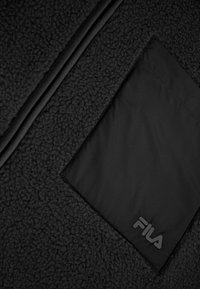 Tela polar negra con una superficie texturizada, que presenta un parche de nylon negro liso. En el parche se encuentra presente un logo tonal de Fila.