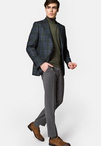 Czarna marynarka w kratę o wyraźnej konstrukcji, oliwkowy golf, szare spodnie i brązowe sznurowane buty. Gładkie faktury i dopasowany krój.