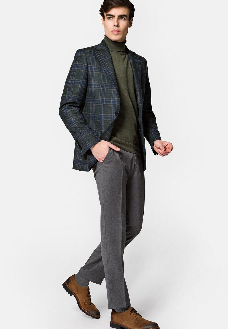 Czarna marynarka w kratę o wyraźnej konstrukcji, oliwkowy golf, szare spodnie i brązowe sznurowane buty. Gładkie faktury i dopasowany krój.