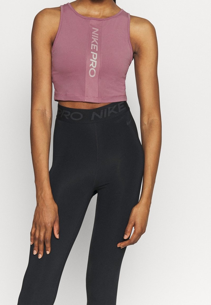 Maui cropowany top sportowy marki Nike Pro w kolorze mauve z odblaskowym pionowym logo, w połączeniu z czarnymi legginsami o wysokim stanie. Gładka tekstura, dopasowany krój.