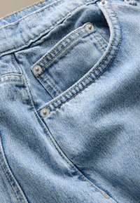 Tissu denim bleu clair montrant la zone de la poche avant avec des coutures orange visibles et des rivets métalliques sur un jean.