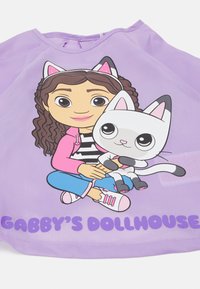 Vijolična majica z risanim junakom s kodrastimi lasmi in črtasto majico, ki drži belo mačko. Na dnu je napis "GABBY'S DOLLHOUSE".