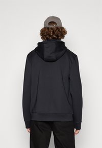 Dark hooded sweatshirt made of smooth fabric, featuring a relaxed fit and cuffed sleeves. Worn with a gray cap. Back view.