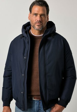 Hombre con cabello corto y oscuro y barba, que lleva una chaqueta con capucha azul marino sobre un suéter marrón y vaqueros azules contra un fondo liso.