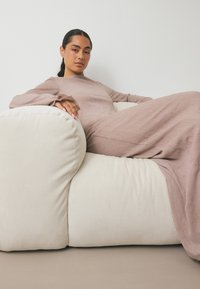 En långärmad, ribbad brun klänning hänger över en texturerad gräddfärgad soffa med rundade hörn, placerad mot en neutral bakgrund.