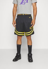 Shorts de basketball noirs avec des accents jaunes et un logo, associés à des chaussettes Nike noires et des baskets montantes de basketball. T-shirt graphique gris.