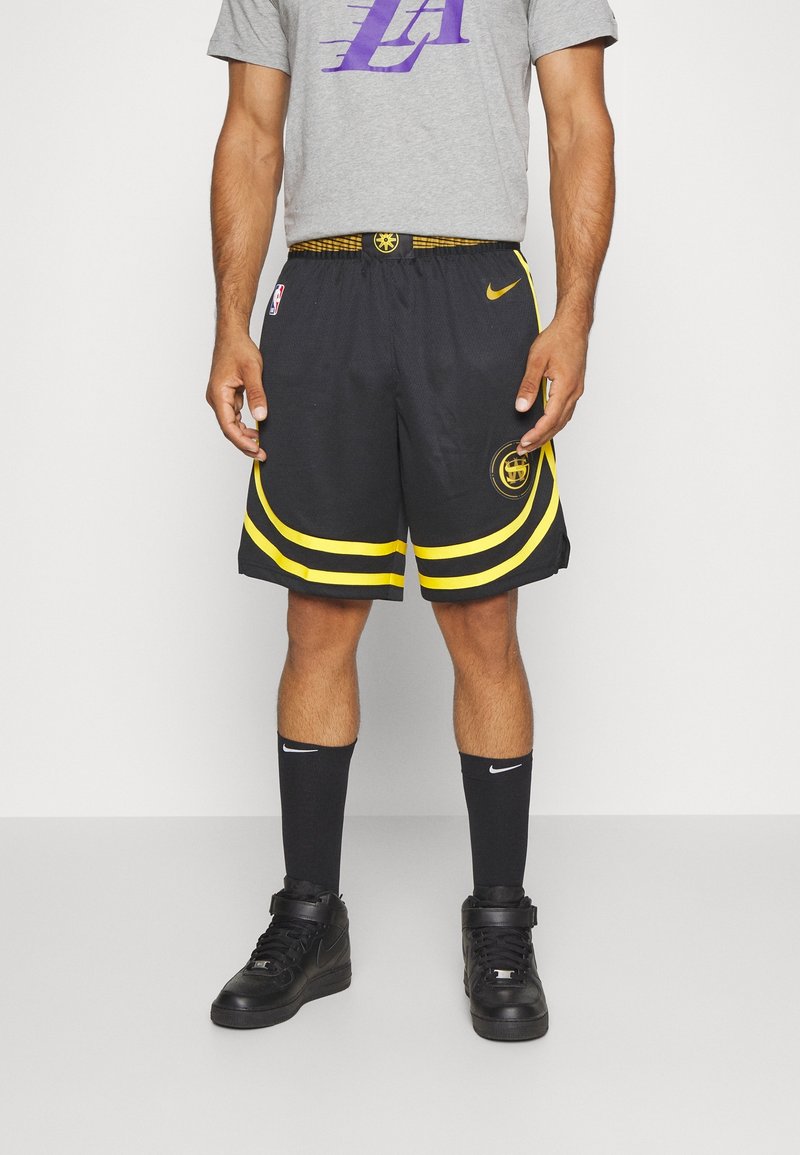Shorts de basketball noirs avec des accents jaunes et un logo, associés à des chaussettes Nike noires et des baskets montantes de basketball. T-shirt graphique gris.