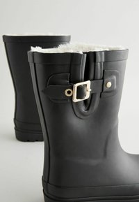 Schwarze Gummistiefel mit einem weißen Kunstfellfutter, ausgestattet mit einer seitlichen Schnallenakzentuierung und einer strukturierten Sohle für besseren Halt. Glatte, matte Oberfläche.