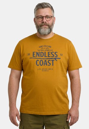 Mężczyzna w średnim wieku z okularami i siwą brodą, ubrany w musztardowożółtą koszulkę z napisem "Endless Coast" oraz oliwkowe spodnie.