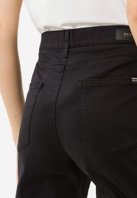 Nahaufnahme einer Person, die schwarze hoch taillierte Hose trägt, mit sichtbarem Bund und Taschen, vor einem verschwommenen hellen Hintergrund.