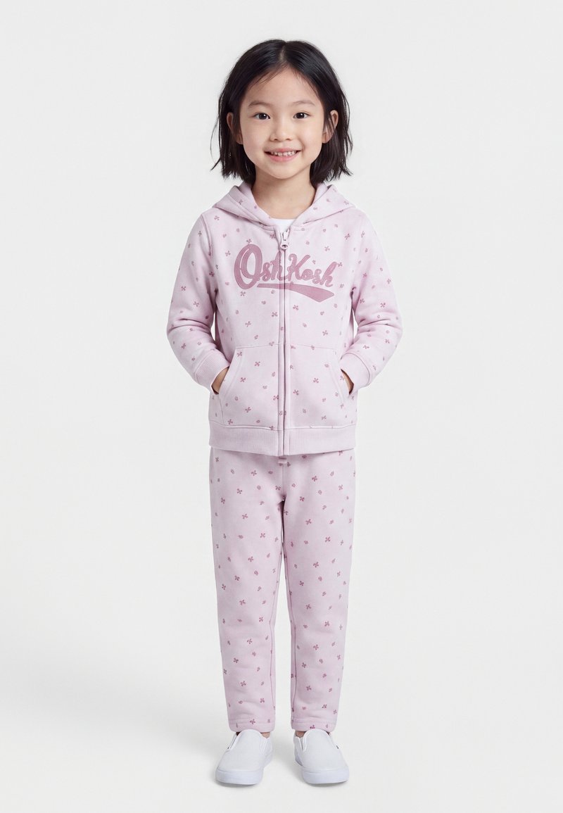 Jeune fille portant un sweat à capuche rose clair et un pantalon assorti avec de petits imprimés floraux, debout et souriante, les mains dans les poches.