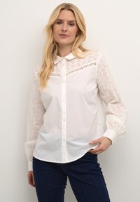 Cămașă albă cu față solidă și model brodat cu ochiuri la mânecile lungi. Gulerul este clasic, iar materialul pare moale și neted.