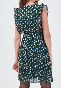 Robe courte plissée en tissu vert foncé, ornée de motifs de feuilles dorées et blanches, avec des manches à volants et une taille cintrée.