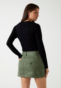 Zwarte lange mouwen top gecombineerd met een olijfgroene rok met een achterzak en subtiel driehoekig logo detail. Over het algemeen gladde texturen.