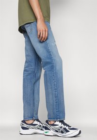 Jeans drepturi de culoare bleu deschis cu un genunchi distrus, asortați cu pantofi sport navy și albi cu accente verzi. Cămașă de culoare verde măsliniu.