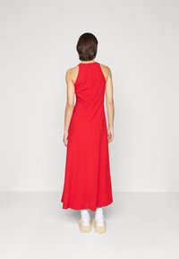 Robe maxi sans manches rouge avec un corsage ajusté et un ourlet évasé, fabriquée en tissu lisse. Le modèle porte des baskets plateforme blanches.