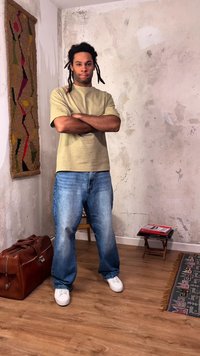 Muž s dredy stojí se založenýma rukama v opotřebované místnosti, má na sobě béžovou košili, modré džíny, bílé tenisky, vedle něj stojí hnědá kožená taška.